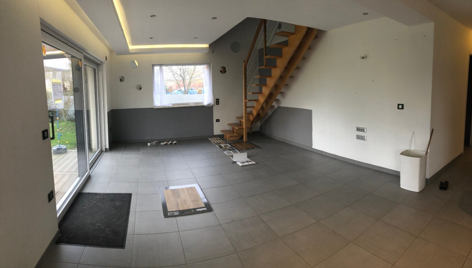 Intérieur d'une maison avec un escalier, sol carrelé et grandes fenêtres.