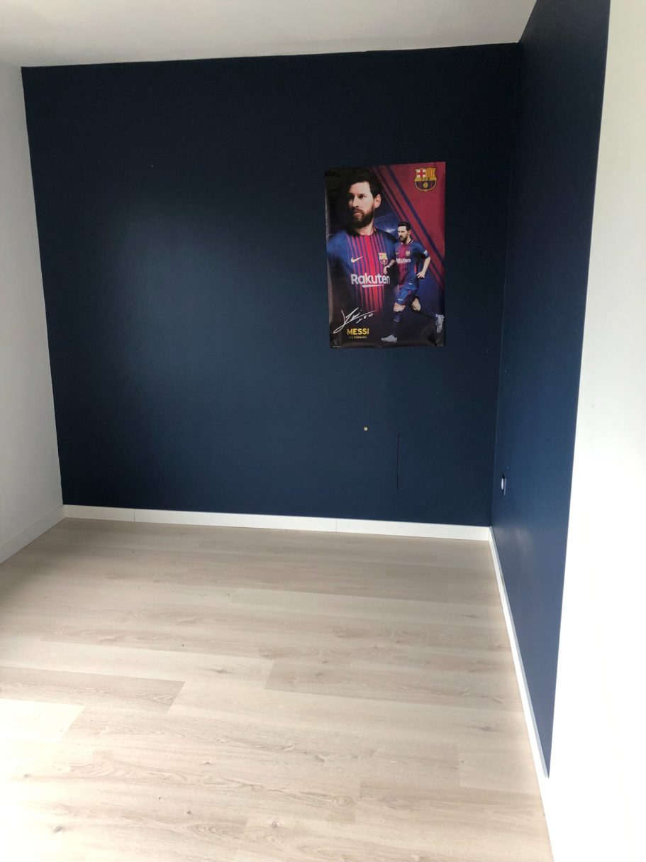 Chambre vide avec un mur bleu et une affiche accrochée. Sol en parquet clair.